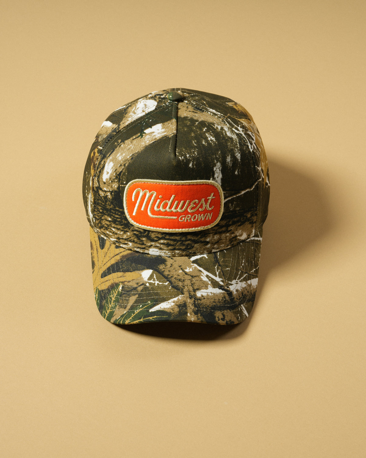 Top view of the Back Forty Camo Hat showing its tree-and-branch print, orange Midwest Grown patch, and brim stitching.