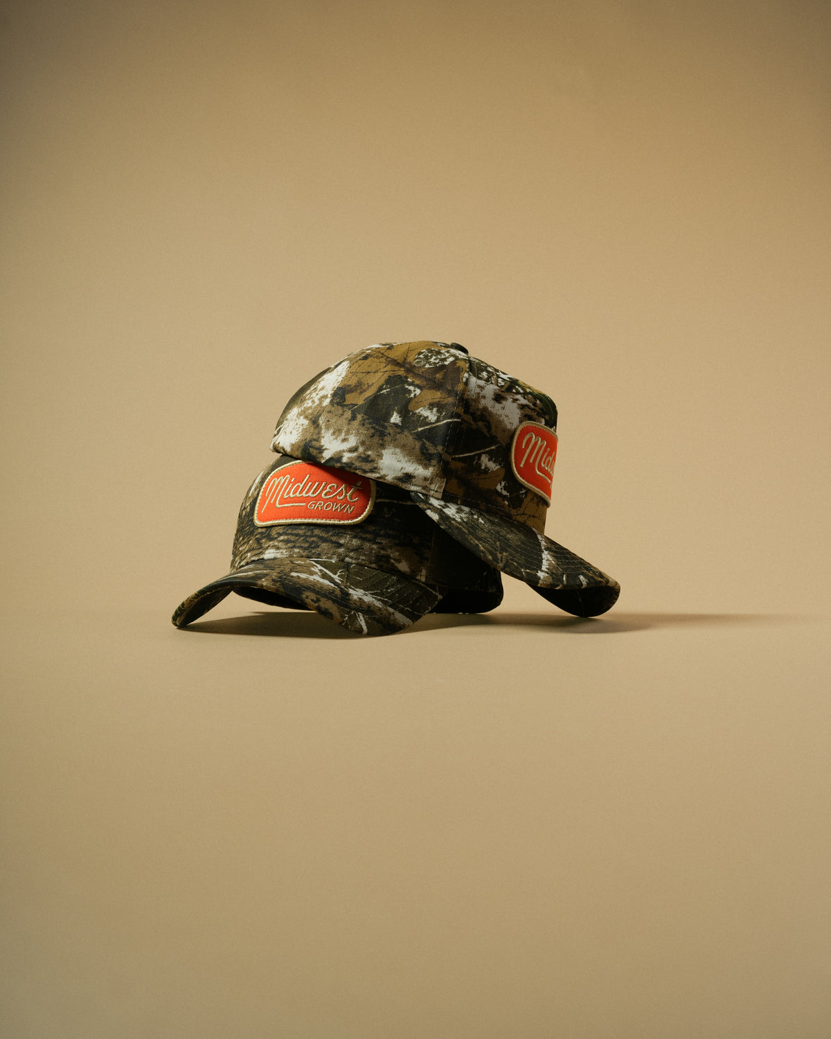 A camouflage baseball cap with two red oval Midwest patches, displayed on a solid beige background.