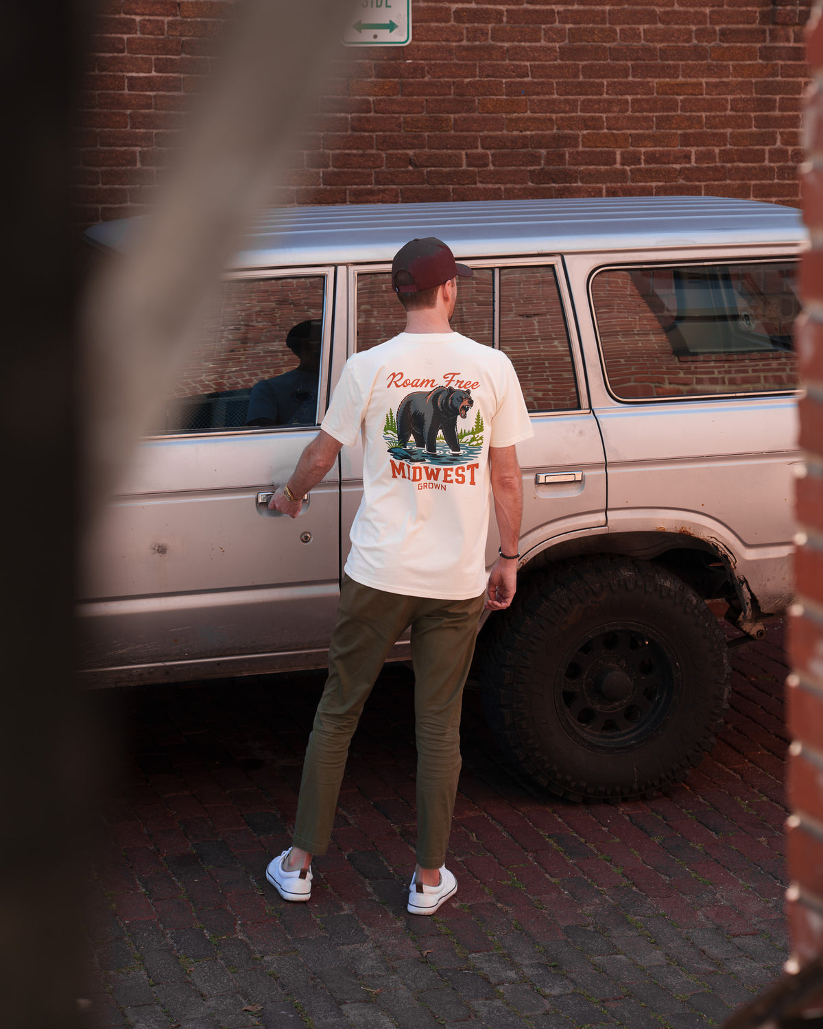 A man stands with his back to the camera next to a silver SUV, wearing a white graphic t-shirt with a 'Midwest' bear design, olive green pants, and white sneakers.