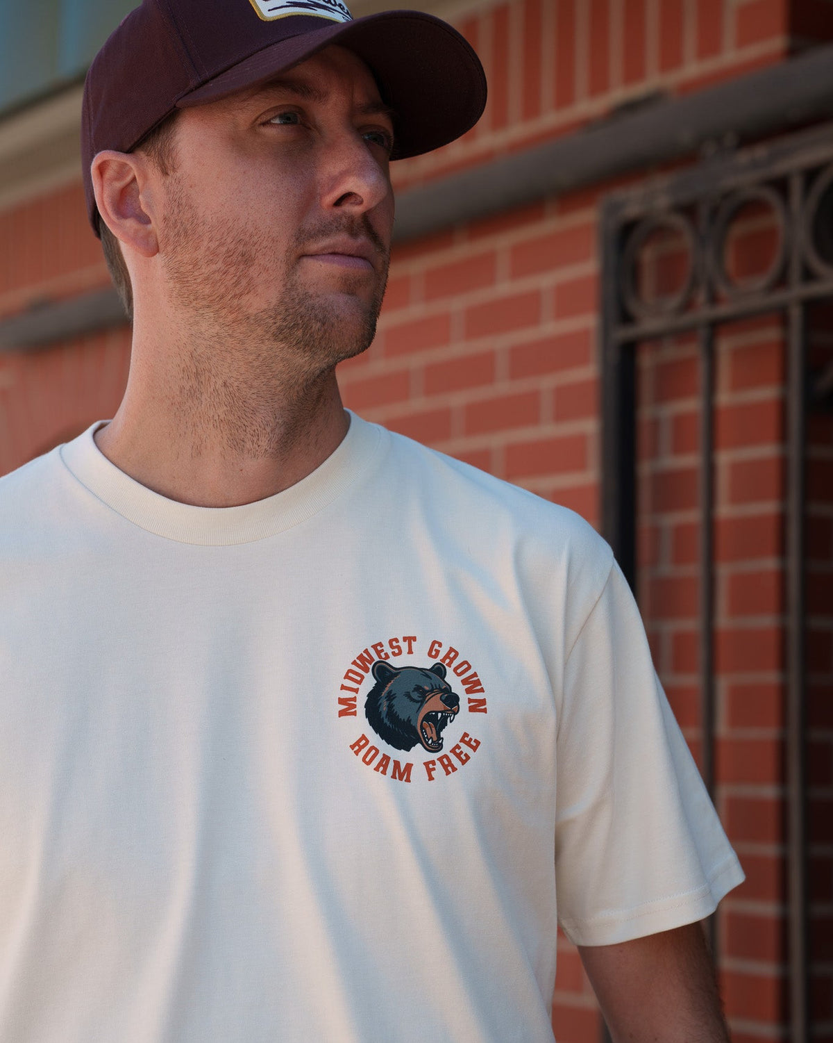 A man wearing a white crewneck t-shirt with a circular 'Midwest Grown Roam Free' graphic on the left chest, paired with a dark maroon baseball cap, standing in front of a red brick wall.