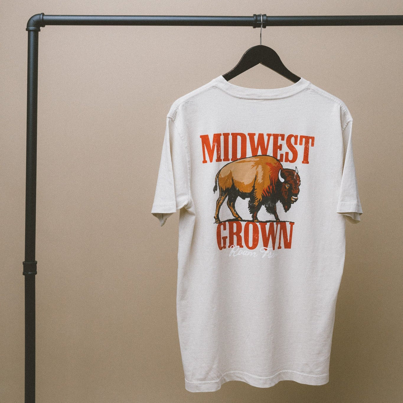 White t-shirt with a graphic of a bison and 'Midwest Grown' text on a hanger against a beige background