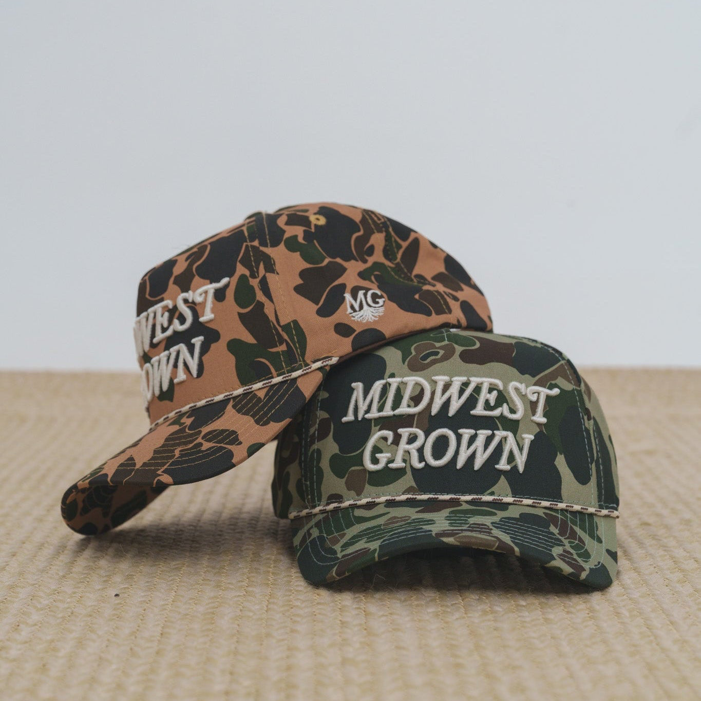 Two duck camo baseball caps with 'Midwest Grown' text on a textured surface.