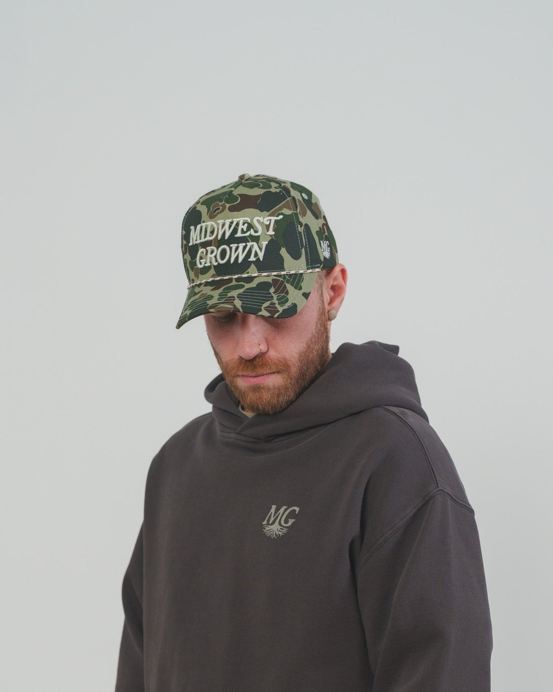 Person wearing a dark Midwest hoodie and camouflage cap with 'Midwest Grown' text on a plain background