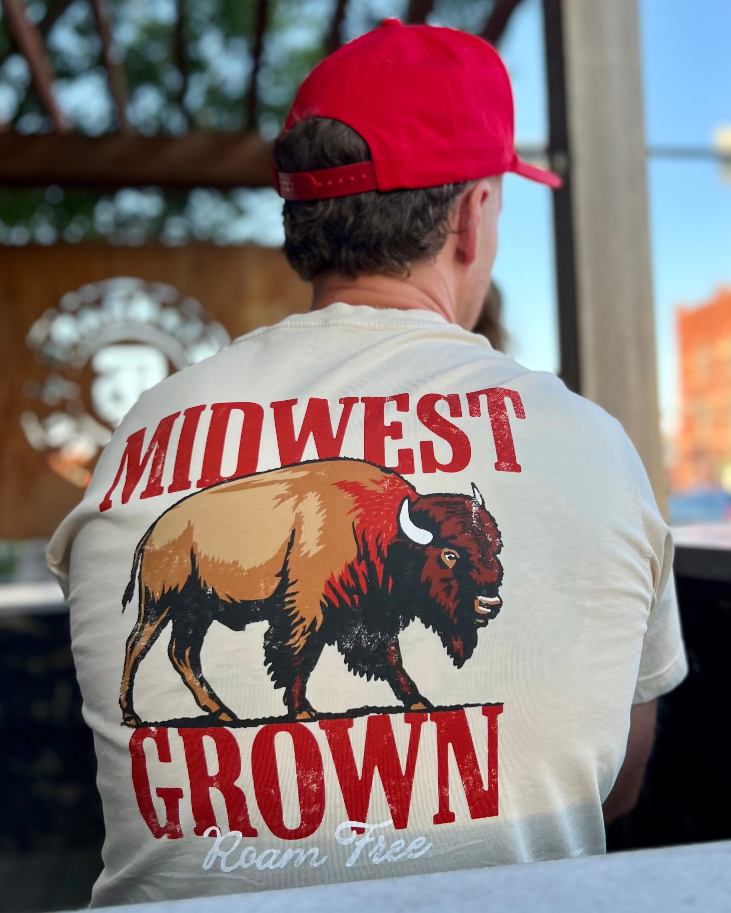 Person wearing a white t-shirt with 'Midwest Grown Roam Free' design and red cap, standing outdoors.