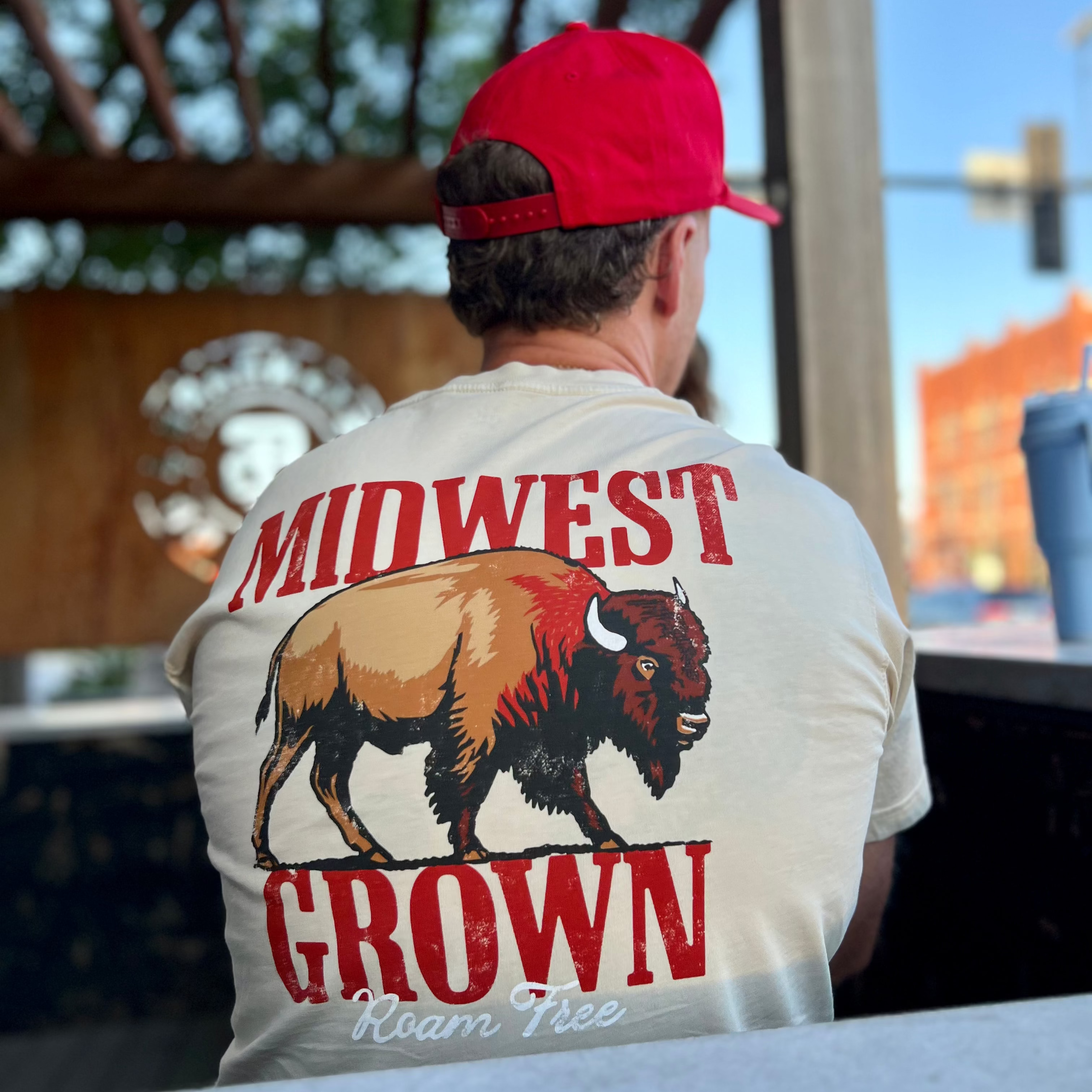 Person wearing a white t-shirt with 'Midwest Grown Roam Free' design and red cap, standing outdoors.