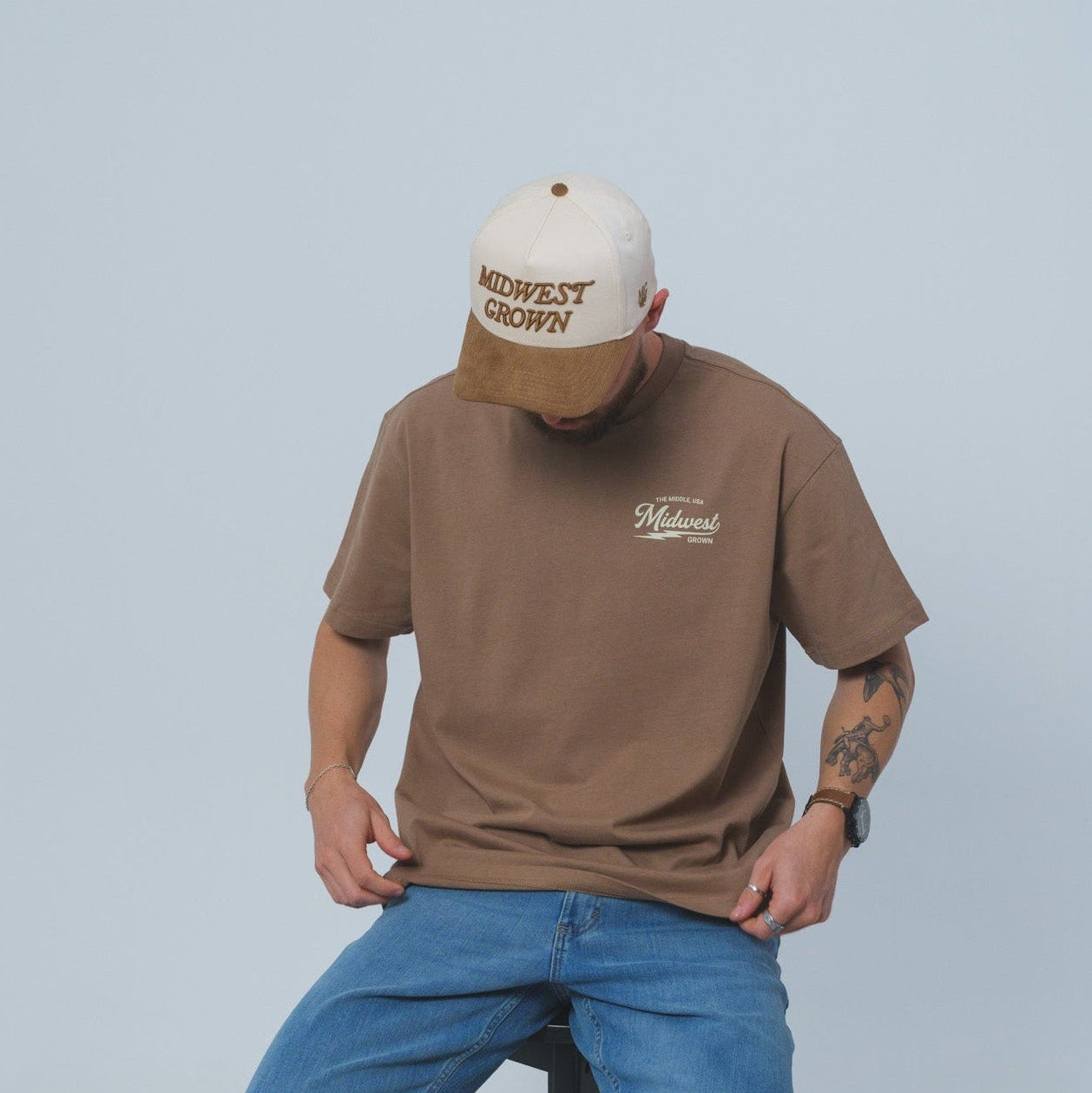 Man wearing a brown Midwest Grown shirt and blue jeans sitting on a stool against a plain background