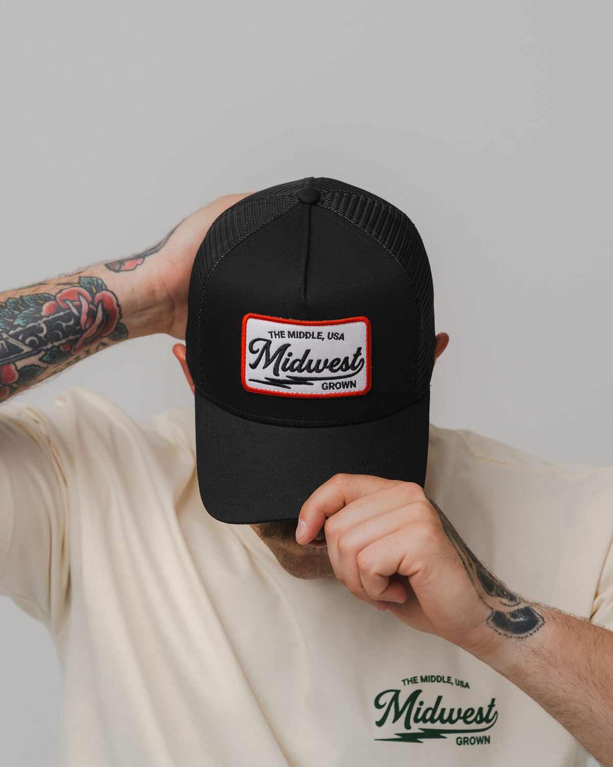 A person wearing a black trucker-style cap with a white and red Midwest Vintage logo patch on the front, paired with a cream-colored t-shirt featuring a matching Midwest Vintage logo on the chest. The person is holding the brim of the cap down with one hand, showing tattooed arms against a solid light gray background.