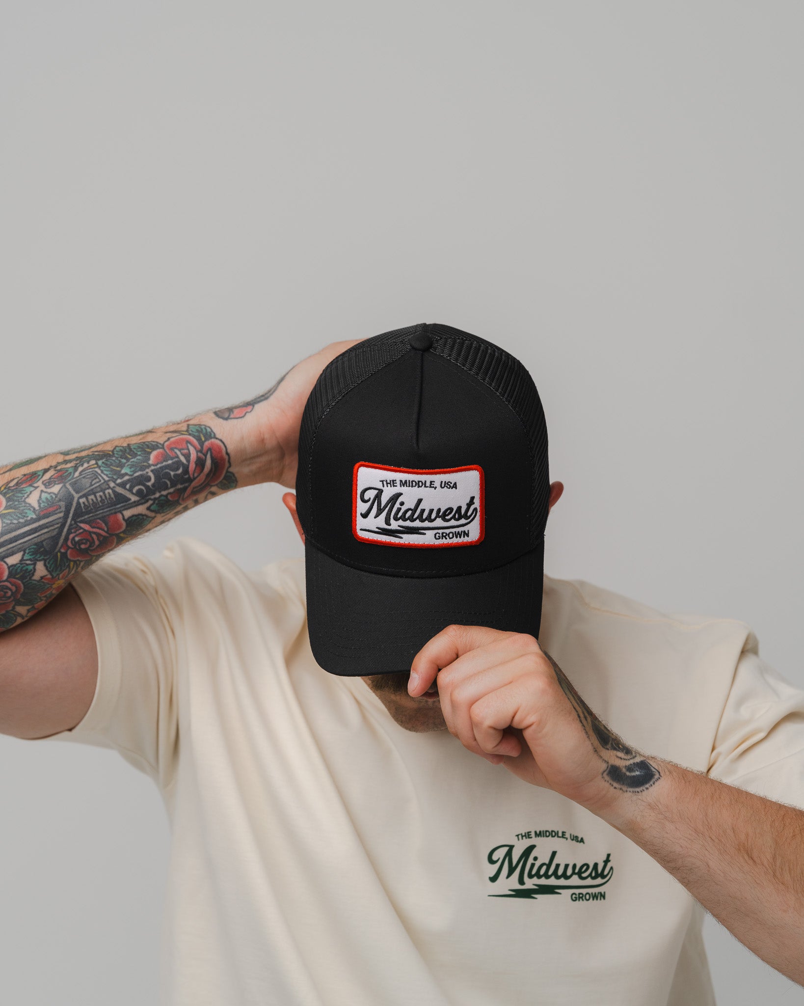 A person wearing a black trucker-style cap with a white and red Midwest Vintage logo patch on the front, paired with a cream-colored t-shirt featuring a matching Midwest Vintage logo on the chest. The person is holding the brim of the cap down with one hand, showing tattooed arms against a solid light gray background.