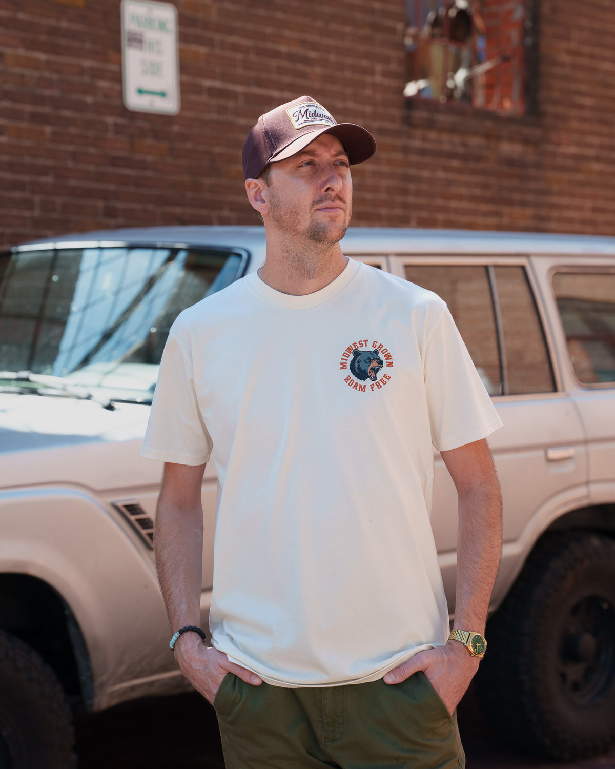 A man wearing a cream-colored t-shirt with a circular 'Midwest Grown' logo on the left chest, paired with a maroon and beige trucker hat and olive green pants, standing outdoors in front of a brick wall and a white vehicle.