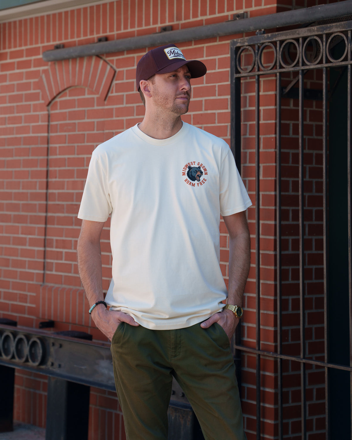 A man wearing a cream-colored t-shirt with a circular graphic on the left chest, olive green pants, and a dark maroon baseball cap, standing in front of a red brick wall. The Tee is a Midwest T-Shirt