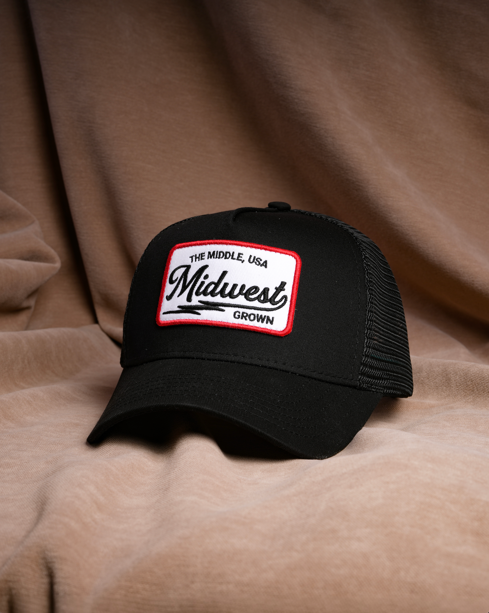 A black trucker-style cap with a white rectangular front patch reading 'The Middle, USA Midwest Grown' in black script and border, photographed against draped tan fabric.