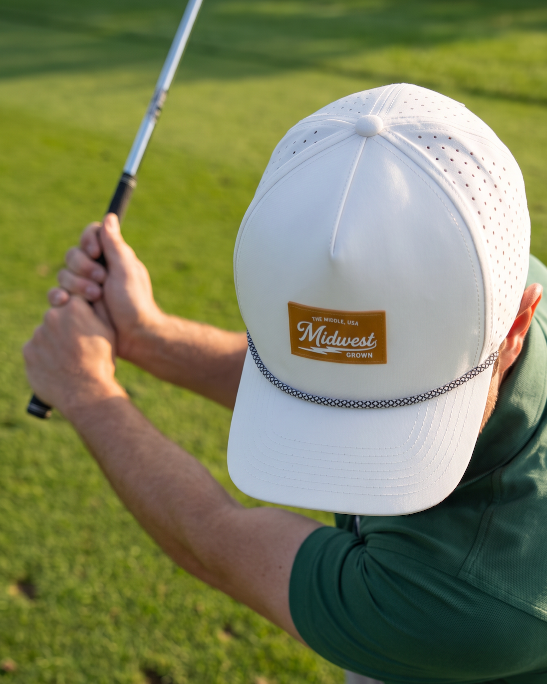 A man on a golf course wearing a white baseball cap with a brown 'Midwest Golf Supply Co.' patch, a green polo shirt, and light pants, captured mid-swing holding a golf club.