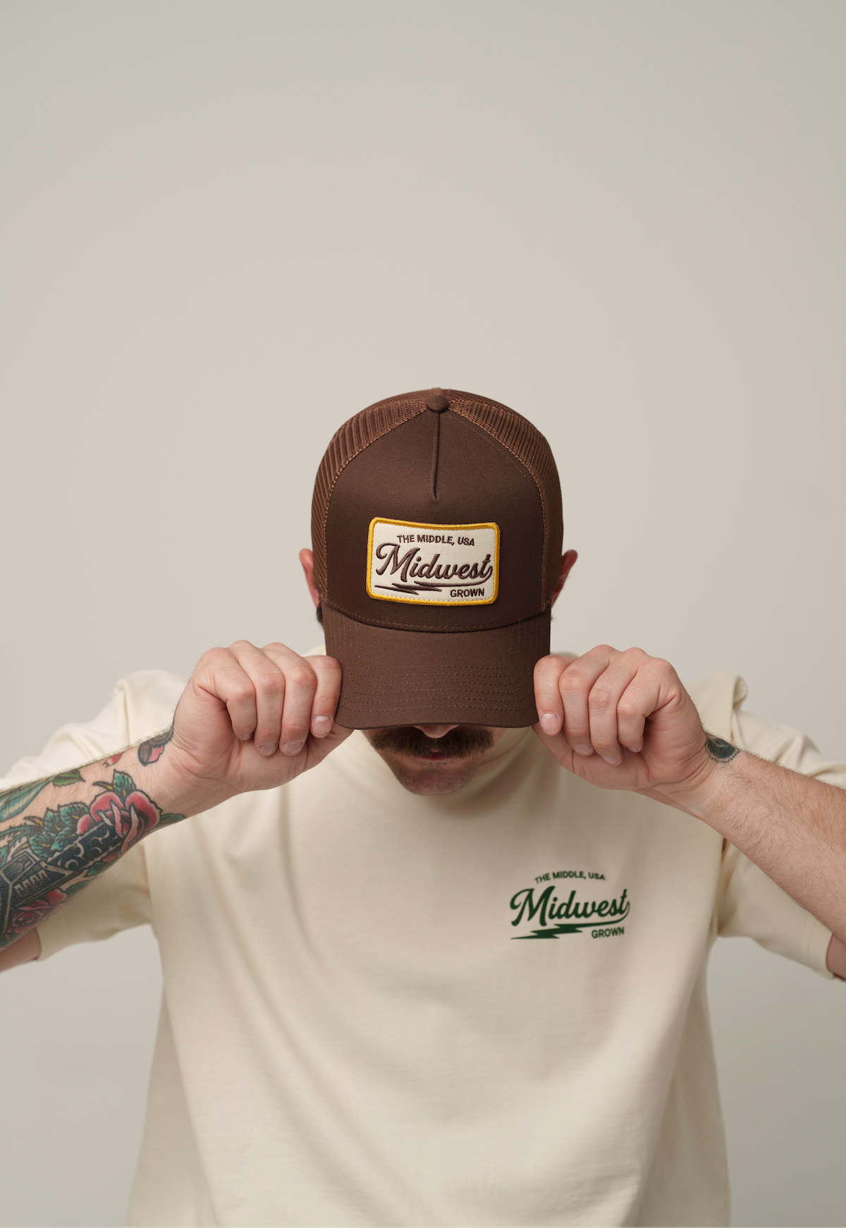 A man wearing a brown trucker hat with a 'Midwest' patch and a cream t-shirt with a matching 'Midwest' logo, holding the brim of the hat in front of his face against a plain light gray background.